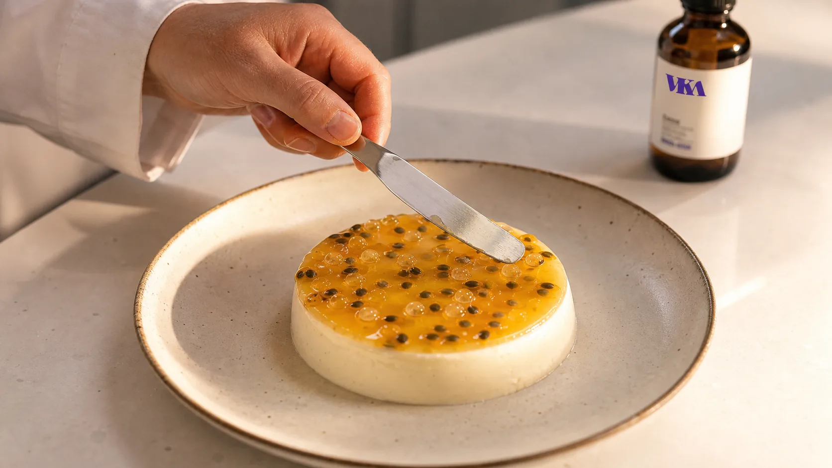 A flavourist's hand on the VKA Australia application bench finishing a small ceramic dessert plate of panna cotta topped with a thin glossy passion-fruit and finger-lime gel, a small offset spatula just lifting away from the plate.