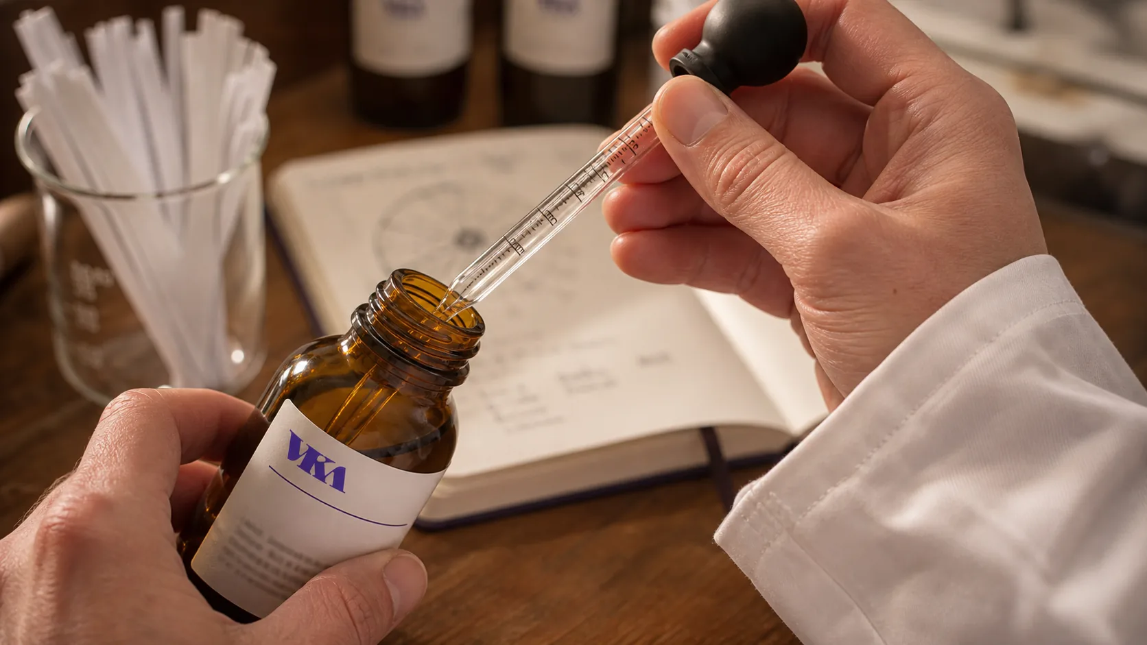 A flavourist pipetting a small dose of essence from an amber bottle into a sample at the VKA Southport compounding bench, white VKA coat with chest mark visible.